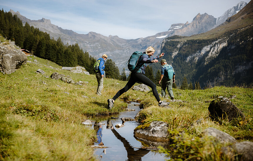 <p>Drei Wanderer überqueren lachend und springend einen Bergfluss.</p>